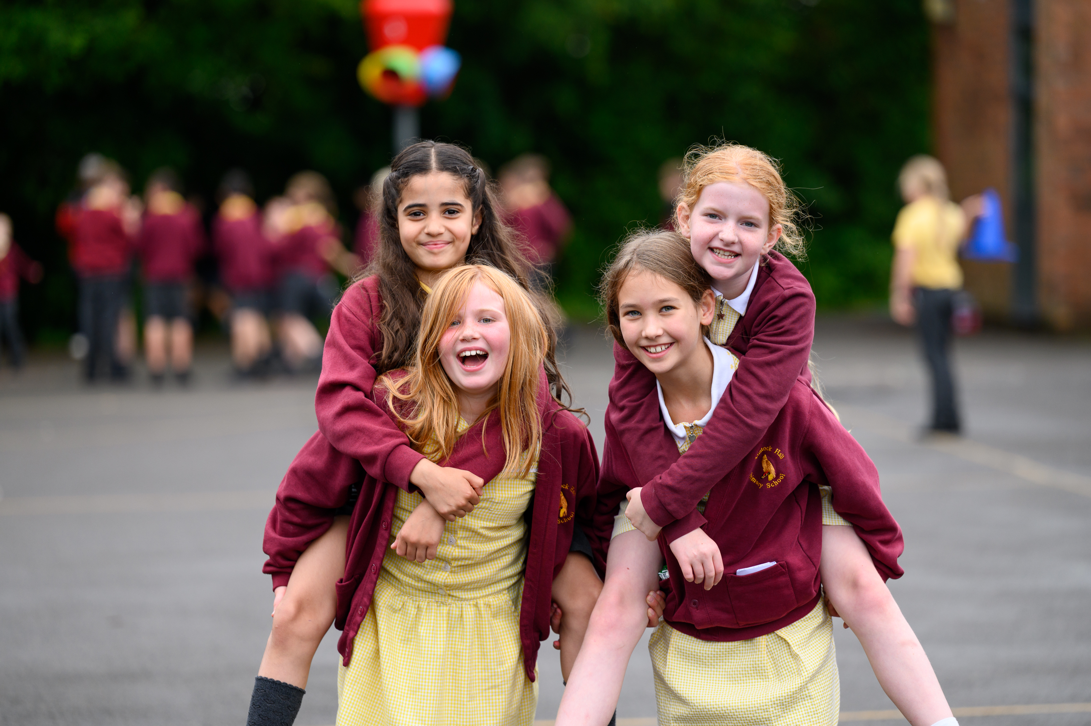 Four students in school uniforms pose together outdoors in a playground, smiling at the camera with other pupils and school buildings visible in the background.