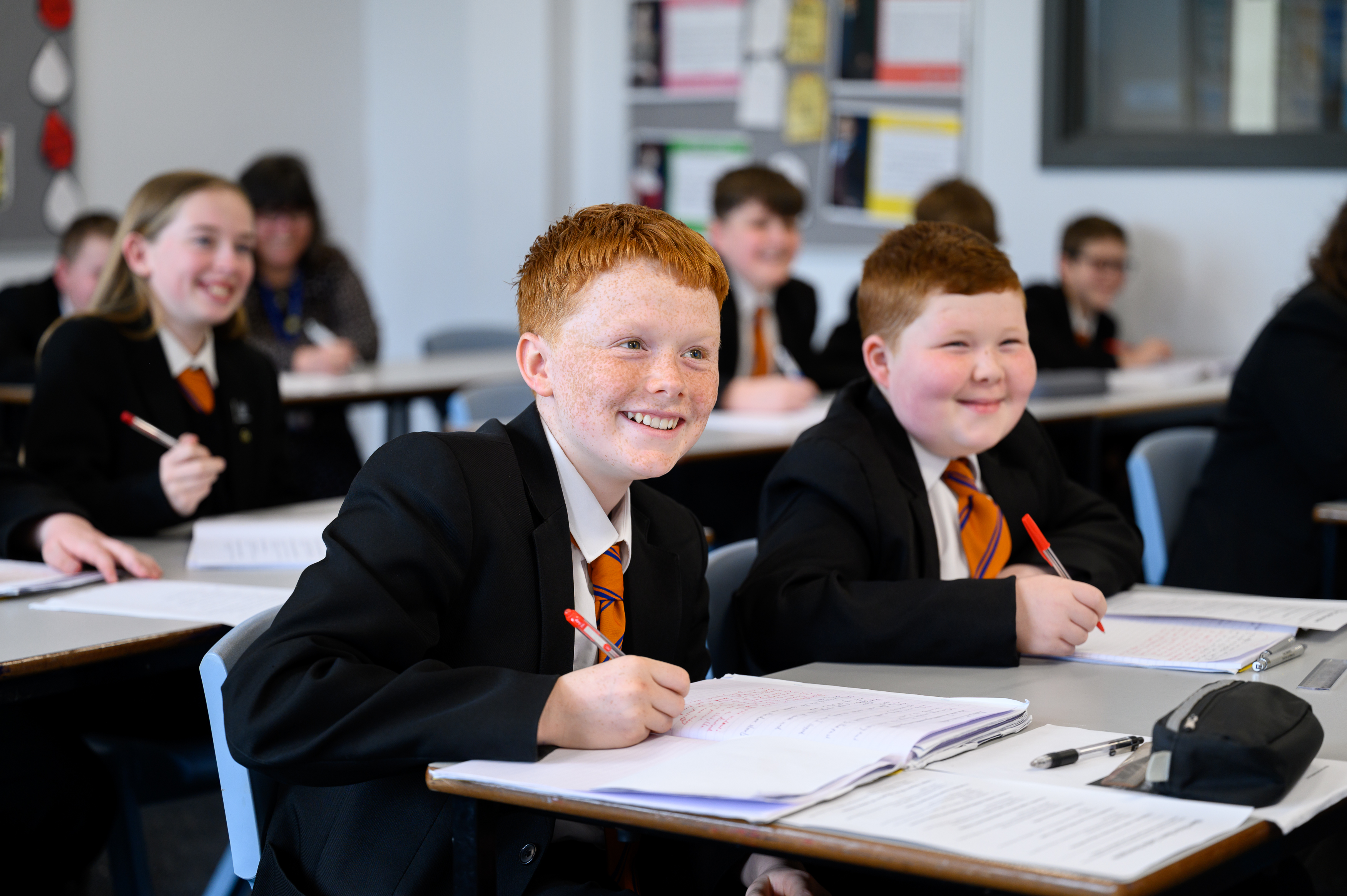 Students in school uniforms sit at desks in a classroom, writing in notebooks during a lesson. Multiple pupils are visible at their workstations with writing materials.