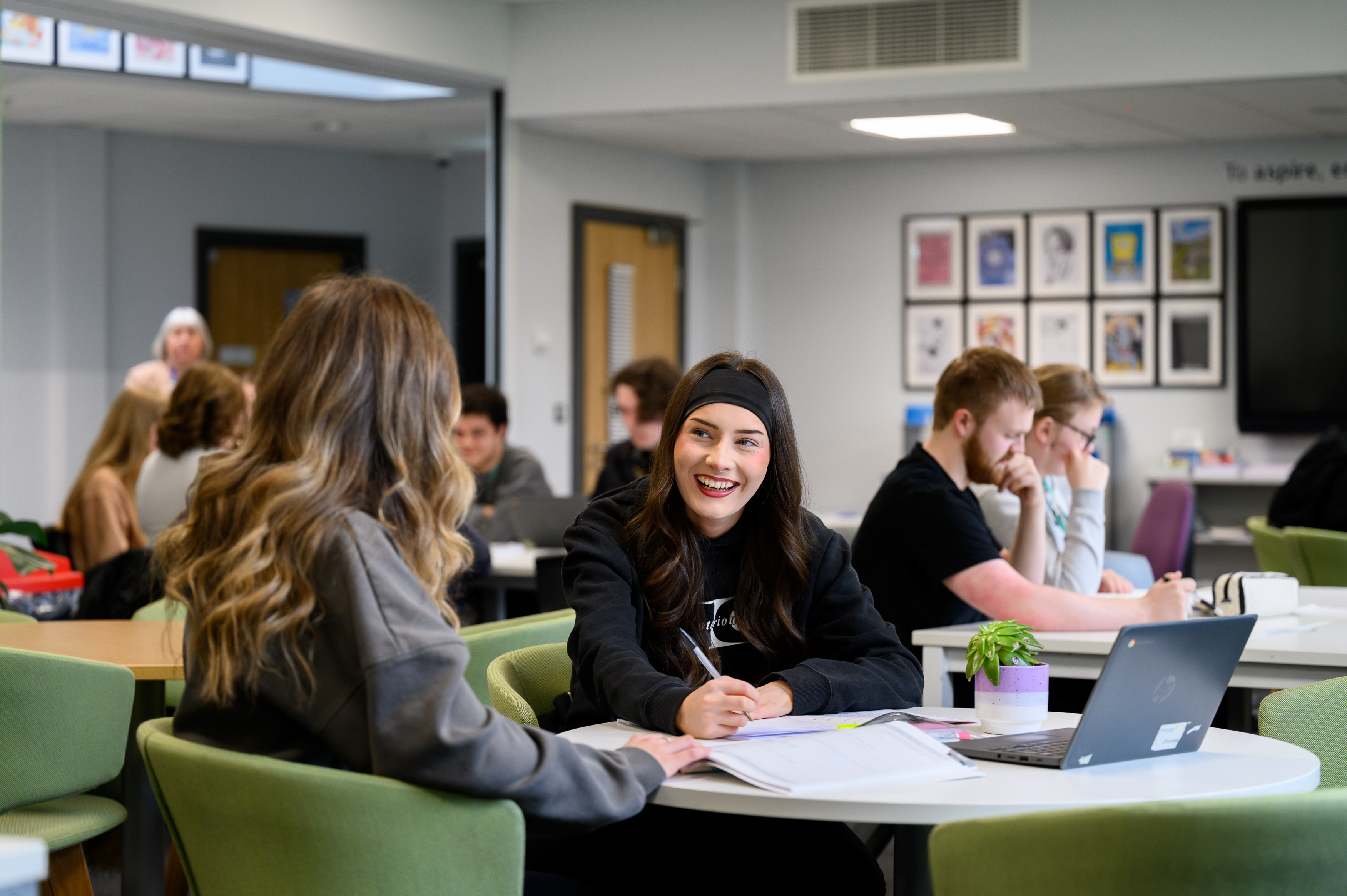 Students work at tables in a modern classroom setting, engaged in individual and collaborative learning activities with laptops and writing materials. Multiple students are seated around white tables with green chairs in a bright, contemporary educational