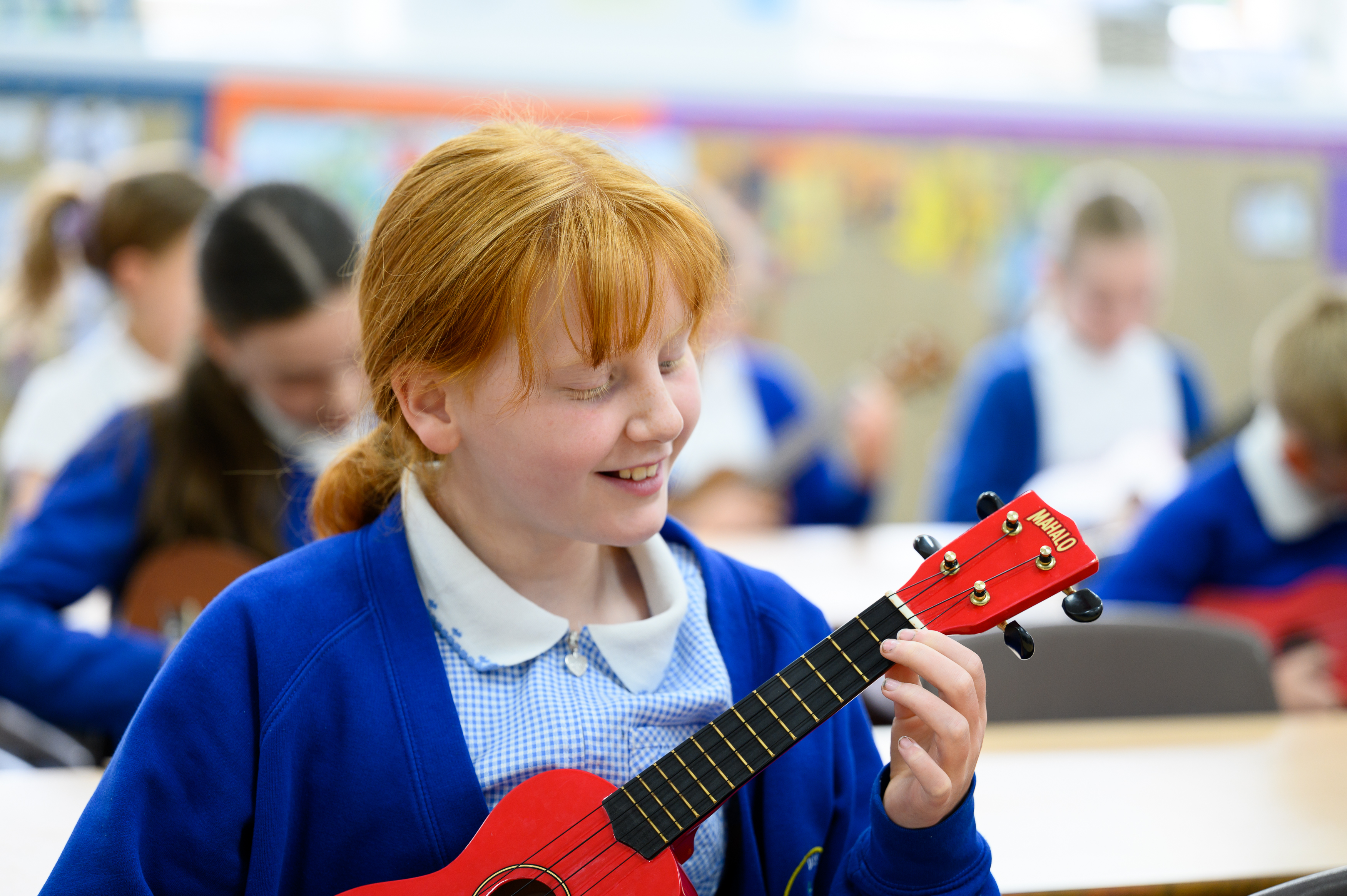 A student in school uniform plays a red ukulele in a classroom setting with other students visible in the background engaged in musical activities.