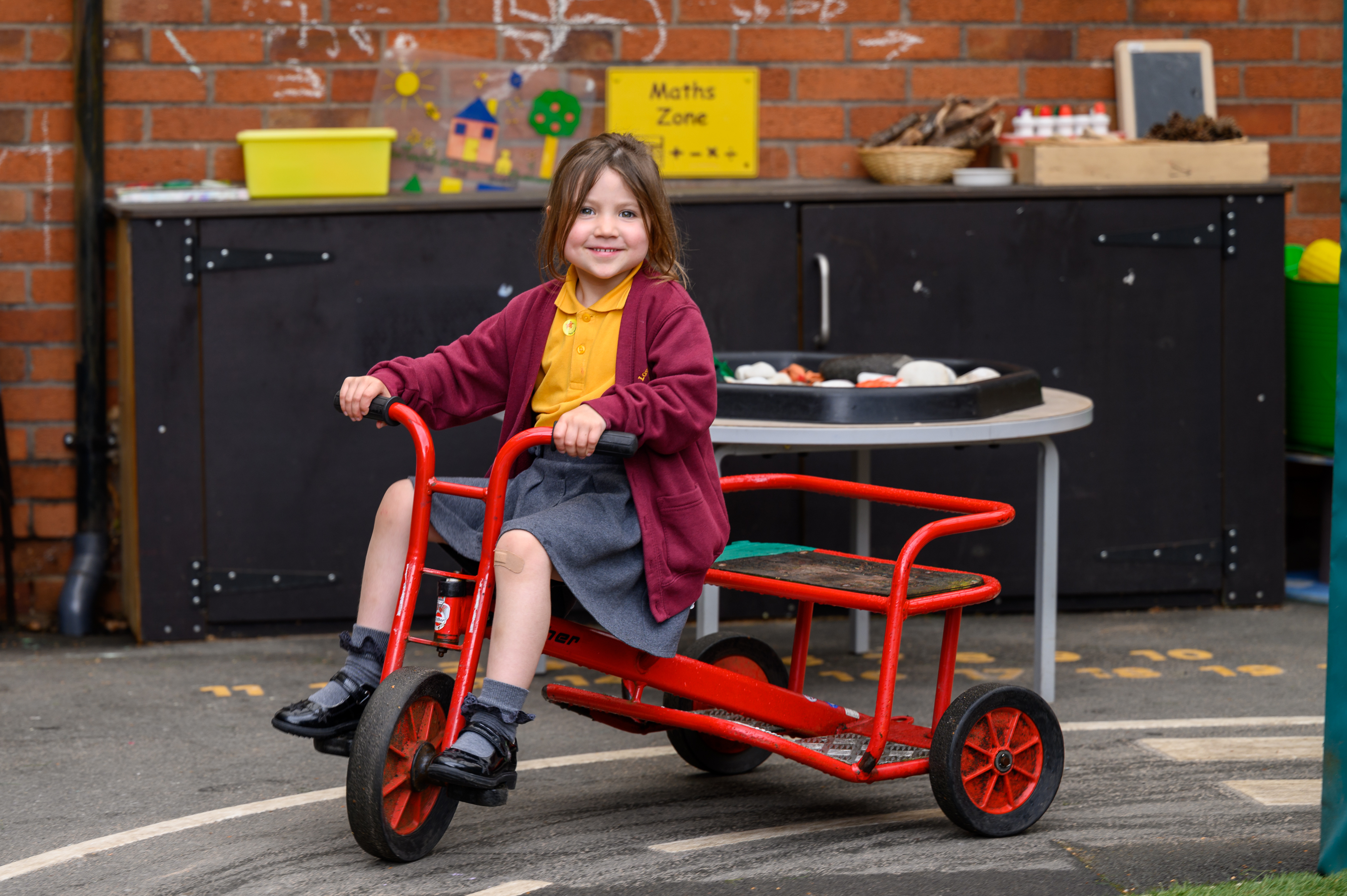 A child rides a red tricycle in a school playground with a brick building and black storage unit in the background.