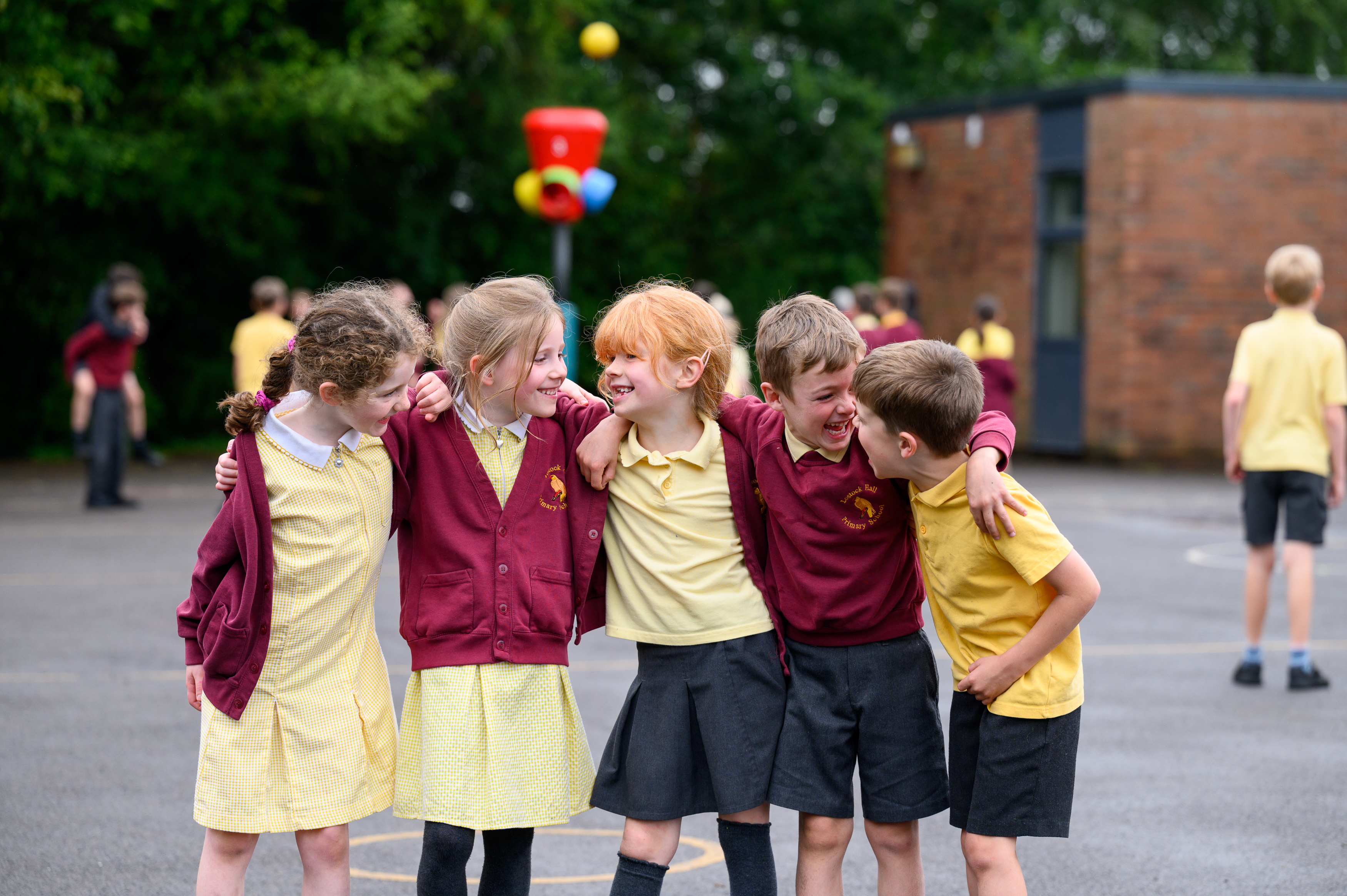 A group of students wearing school uniforms stand together on a playground with a brick building and sports equipment visible in the background during an outdoor school activity.