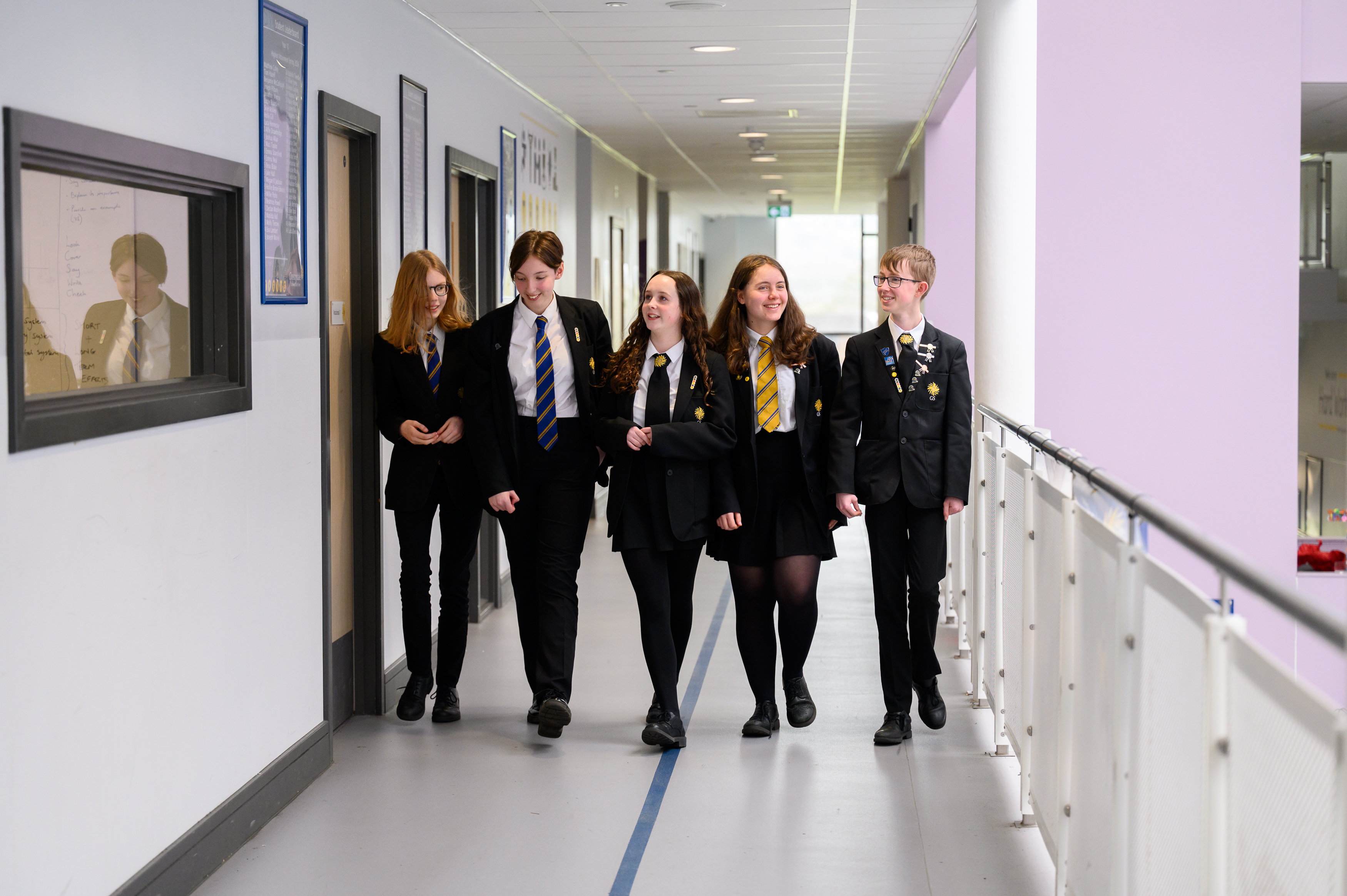 A group of students in school uniforms walk together down a bright, modern school corridor with white walls and purple accents, passing classroom doors and educational displays.