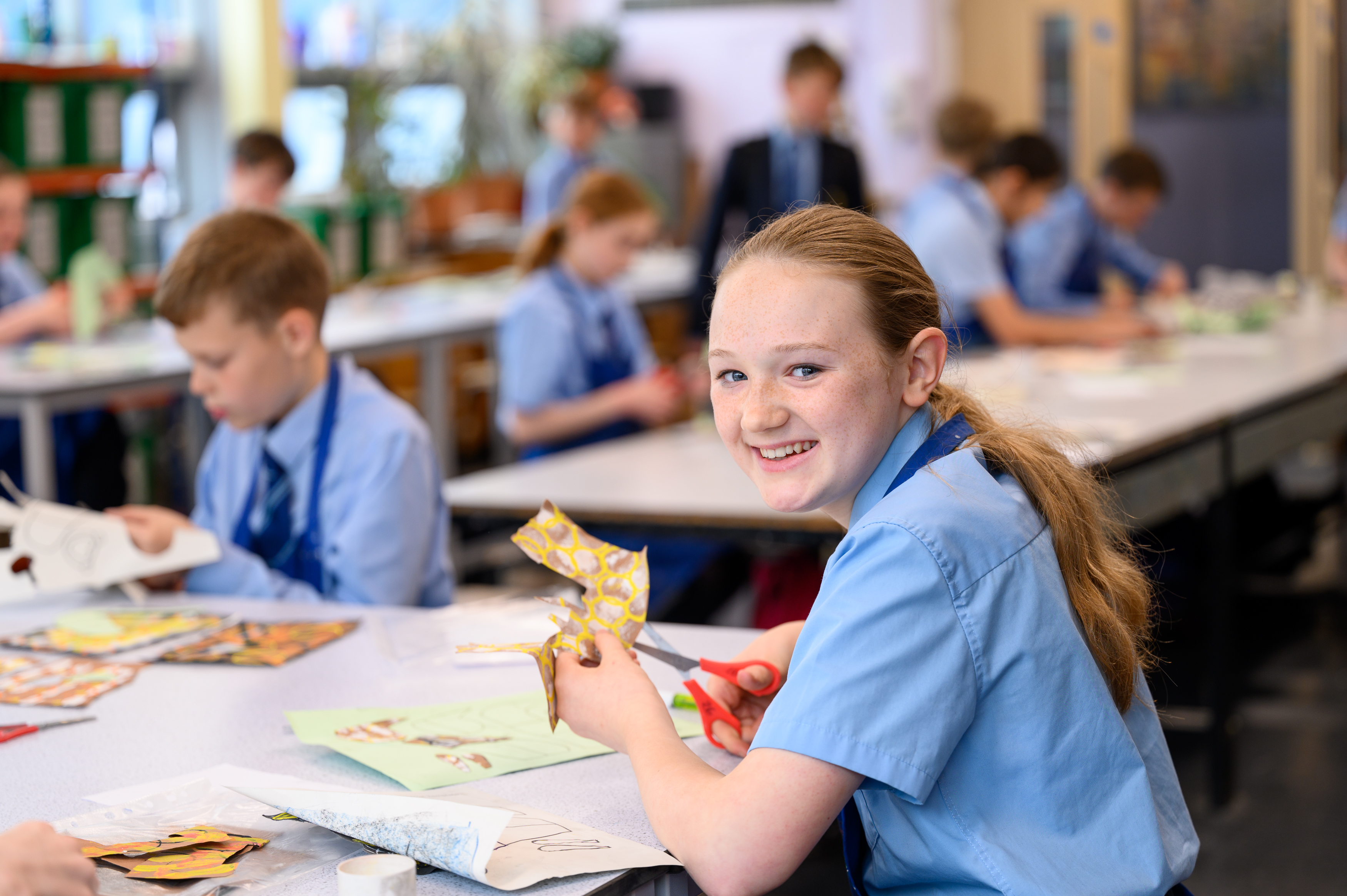 Students in school uniforms participate in an art or craft activity at tables in a classroom, with one student smiling at the camera while working on a colorful paper project.