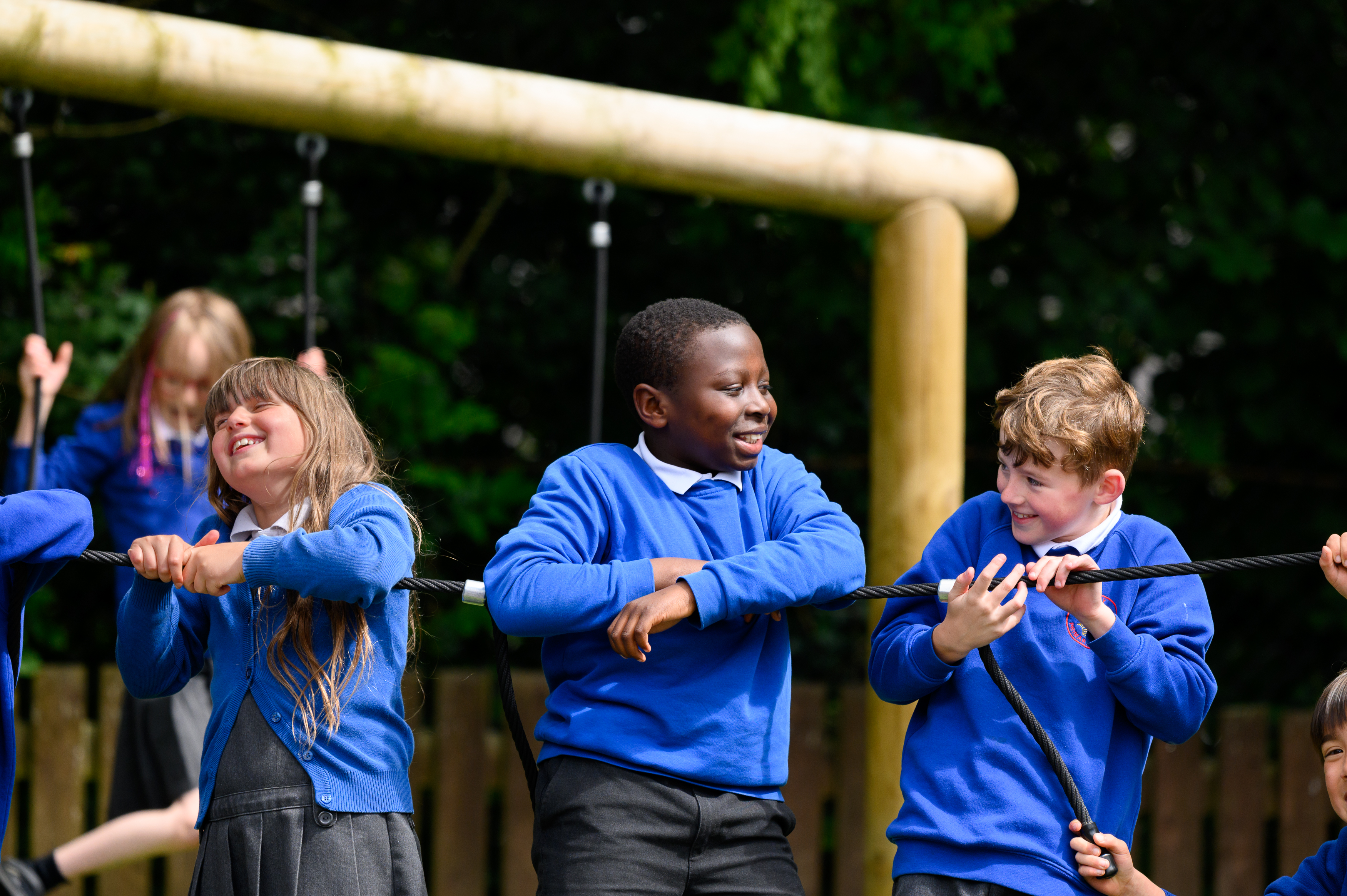 A group of students in blue sweatshirts participate in a tug-of-war activity outdoors, holding a rope together as part of a team-building exercise near playground equipment.