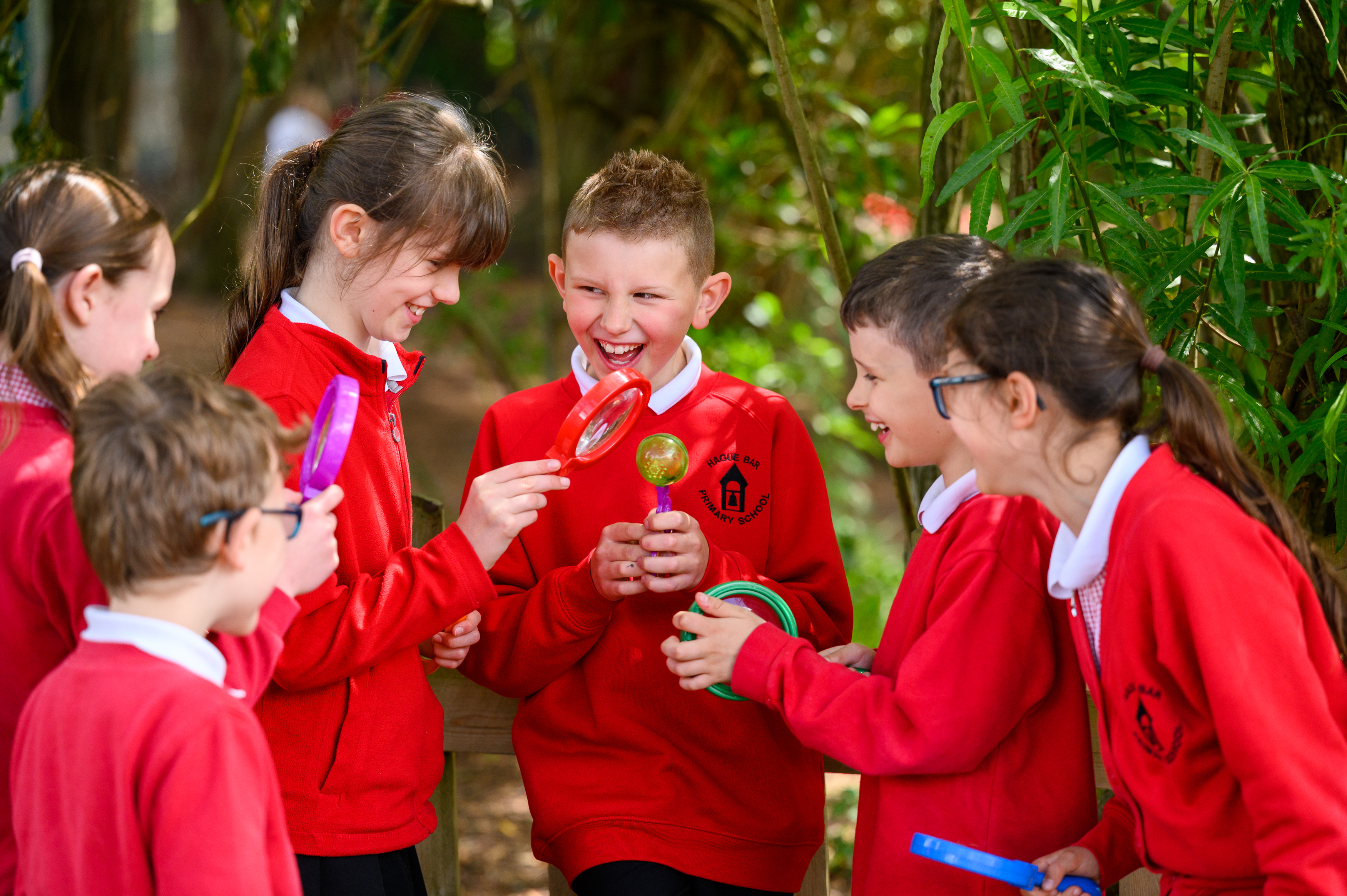 A group of pupils in red school uniforms gather outdoors among green foliage, examining and discussing what appears to be natural specimens or objects during an educational activity in a garden setting.