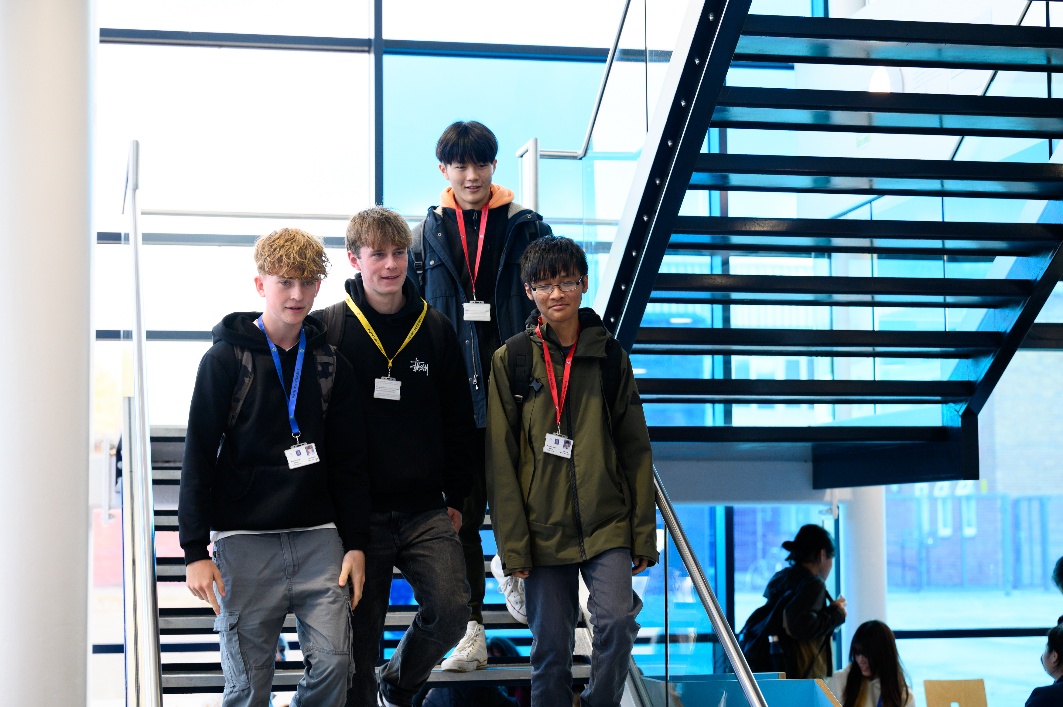 Four young students wearing lanyards with ID badges stand together on a modern indoor staircase with large windows and blue architectural features in the background, appearing to be on a school visit or educational outing.