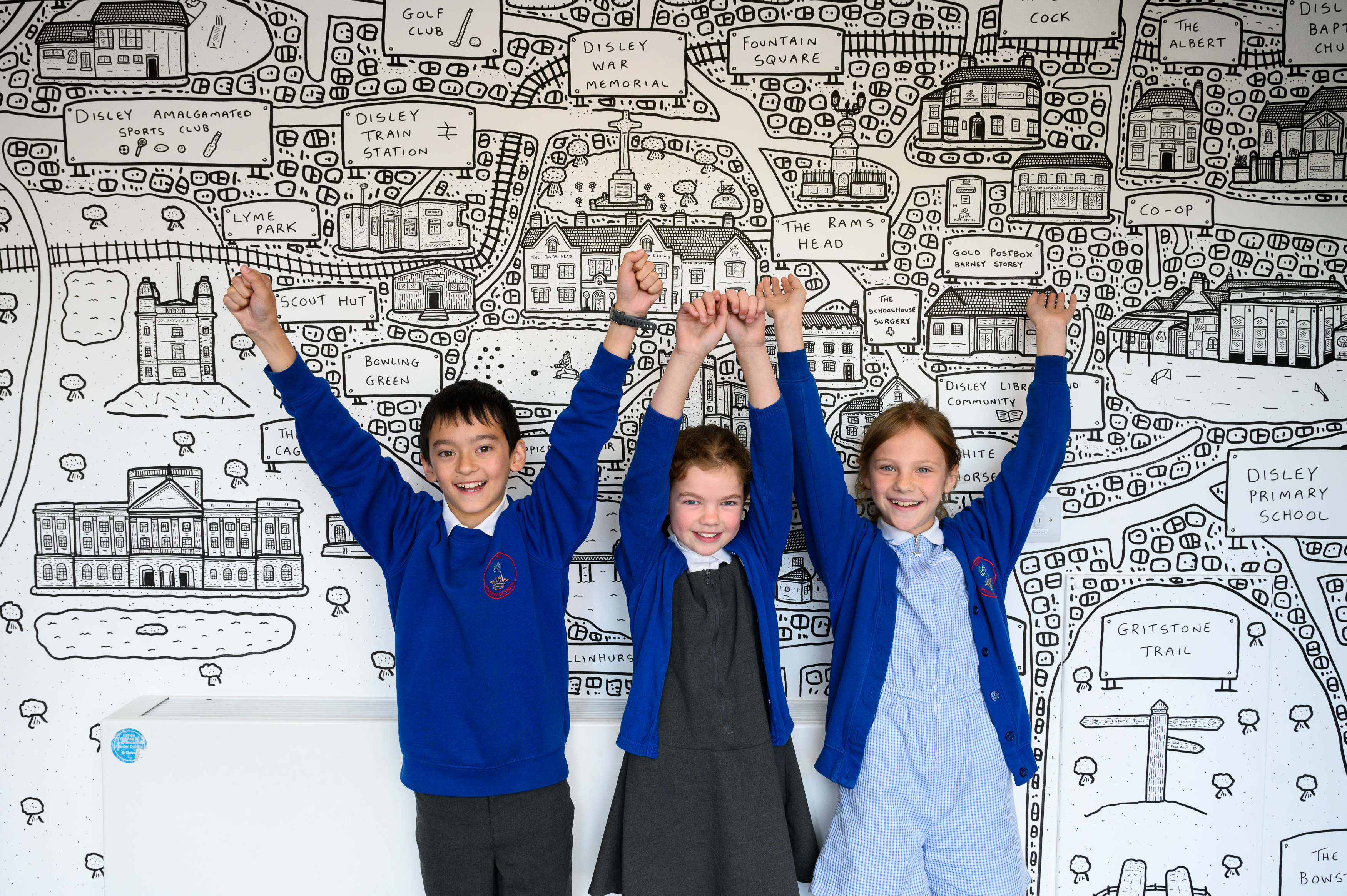 Three pupils in school uniforms stand with arms raised in celebration in front of a large detailed hand-drawn map illustration displayed on a wall, appearing to be part of a classroom art or geography project.