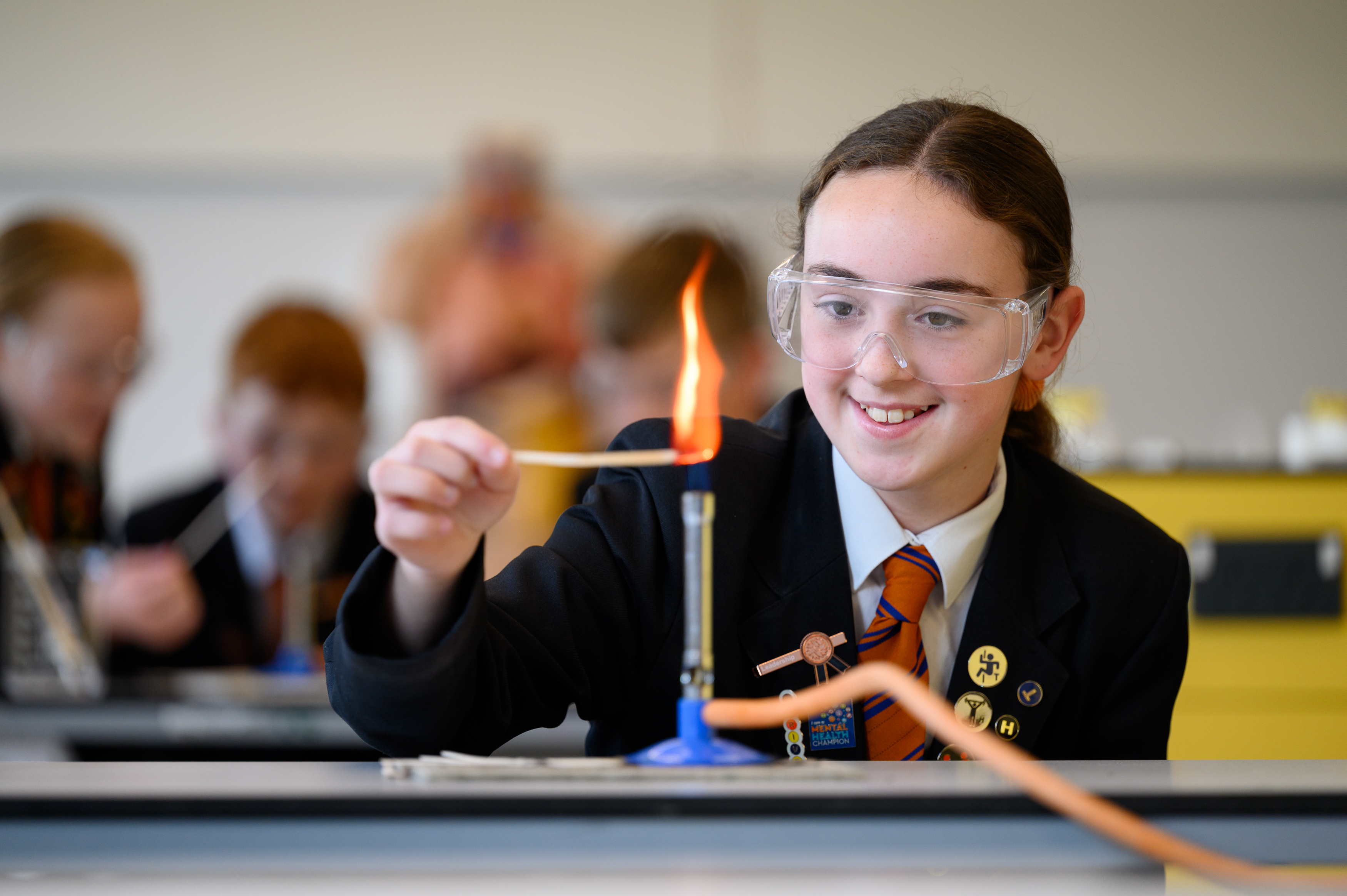 A student wearing safety goggles conducts a chemistry experiment with a lit matchstick near a flame in a science laboratory, with classmates observing in the background.