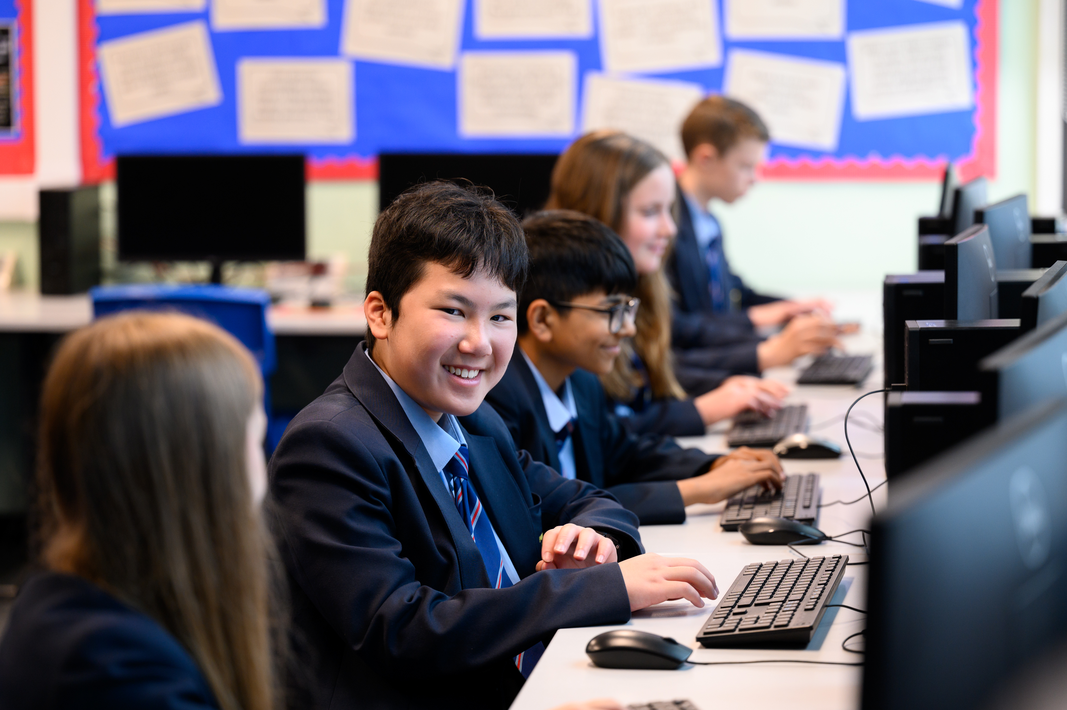 Students in school uniforms work at desktop computers in a classroom, seated at desks with monitors and keyboards. A bulletin board with blue and beige panels is visible on the wall behind them.