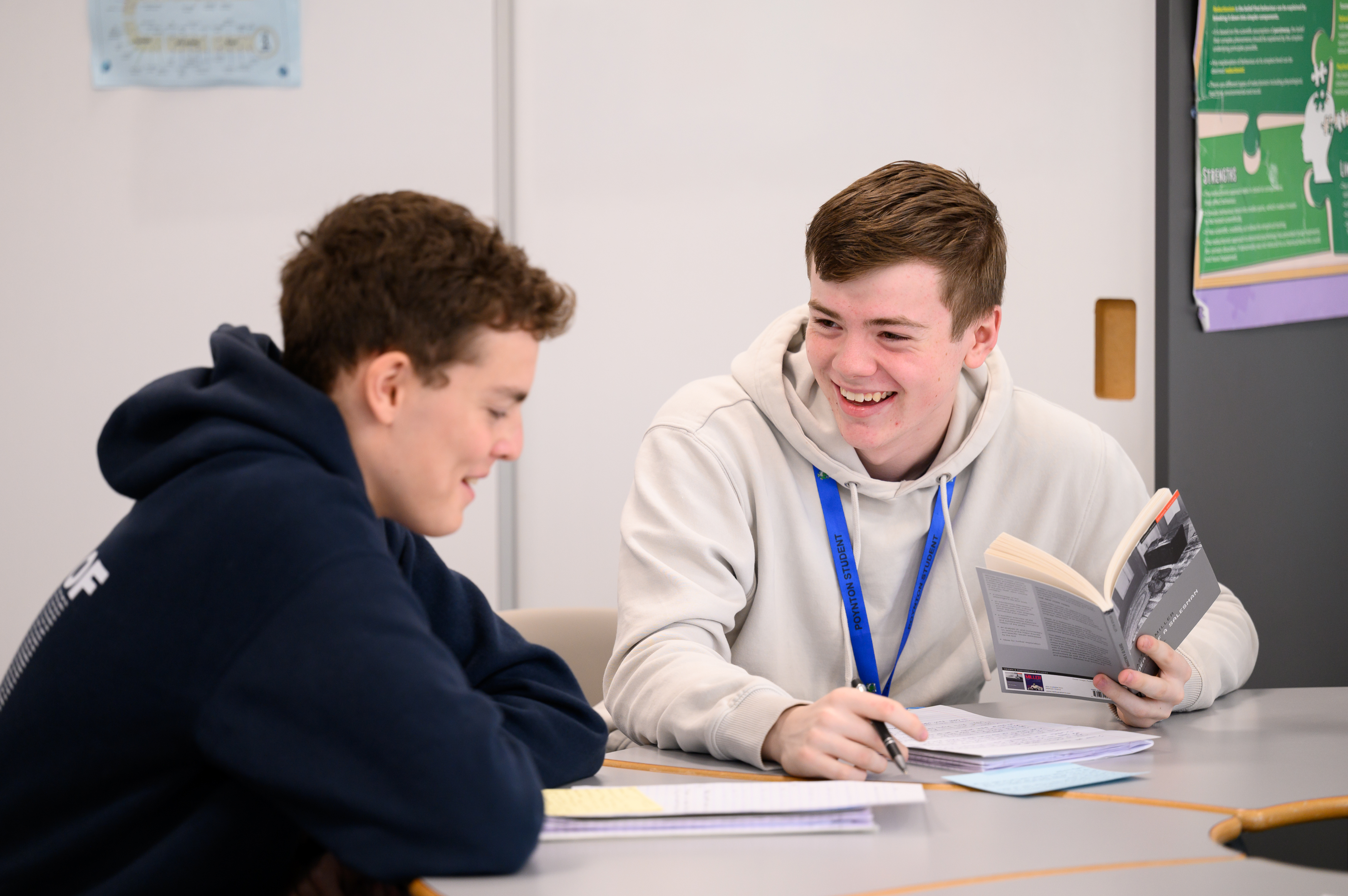 Two students sit at a table in a classroom during a reading session. One student wears a blue hoodie while the other wears a light gray hoodie with a blue lanyard, both engaged with books and notes.