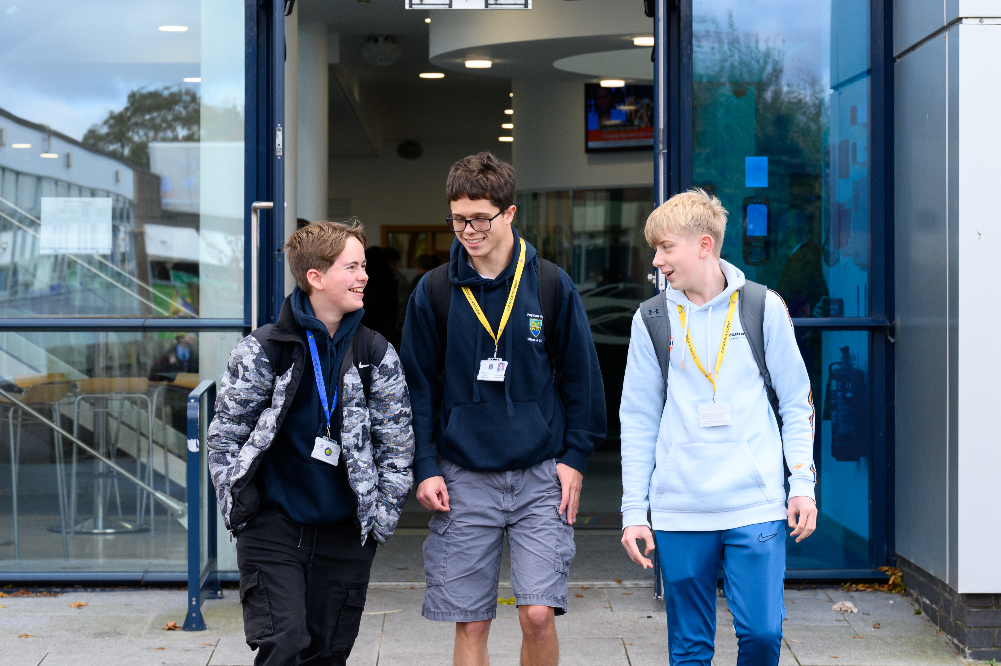 Three students wearing lanyards walk together outside a modern school building with glass doors and windows, appearing to be on a school trip or educational visit.